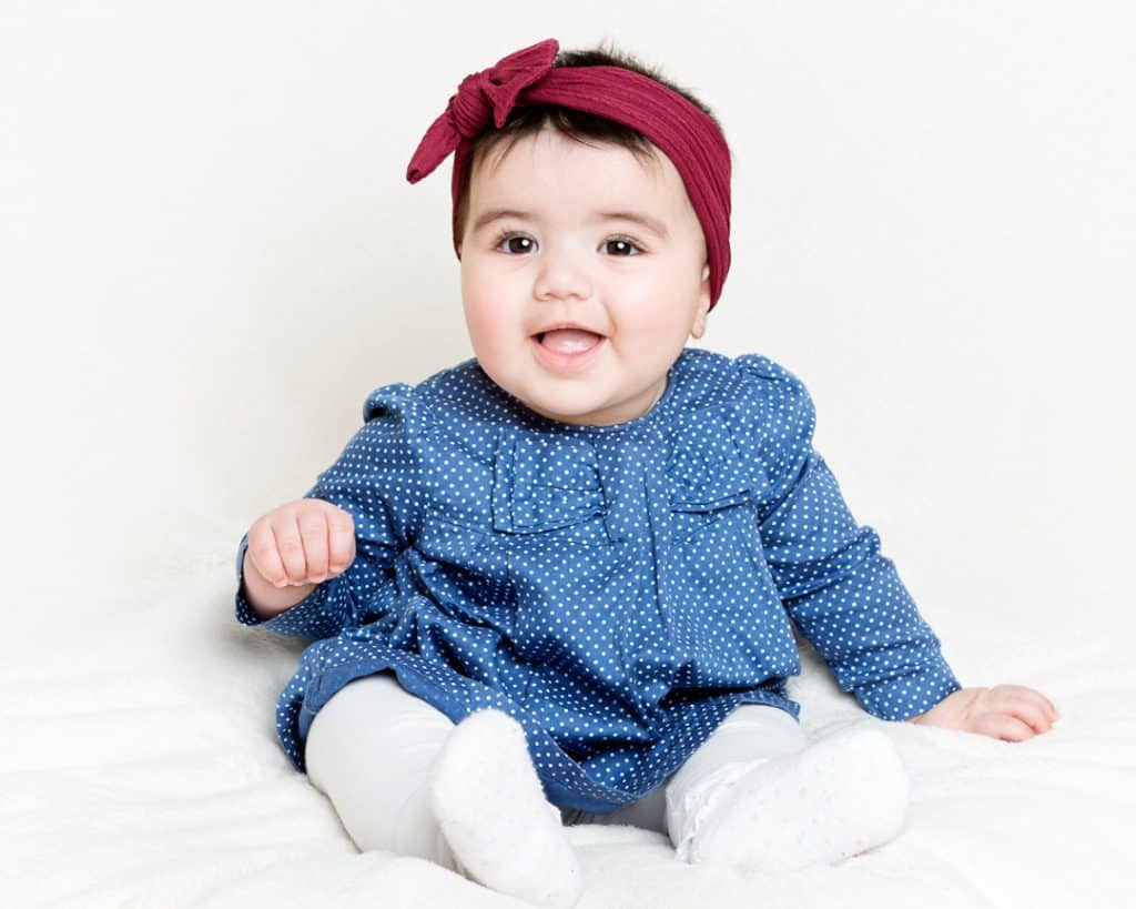 a little girl in a blue dress with a red headband sits on a white blanket