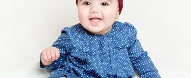 a little girl in a blue dress with a red headband sits on a white blanket