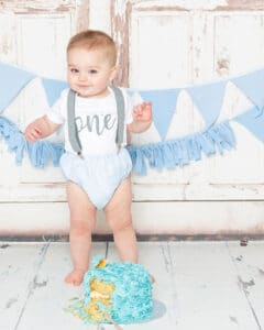 Cake Smash Photography East Grinstead West Sussex a little boy in a vest and bloomers stands behind his blue cake at a cake smash photo shoot