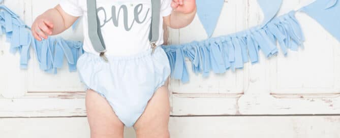a little boy in a vest and bloomers stands behind his blue cake at a cake smash photo shoot