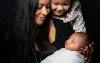 a family of a mother and her older daughter look down at a sleeping newborn wrapped in a cream swaddle