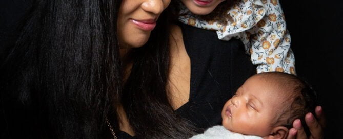 a family of a mother and her older daughter look down at a sleeping newborn wrapped in a cream swaddle