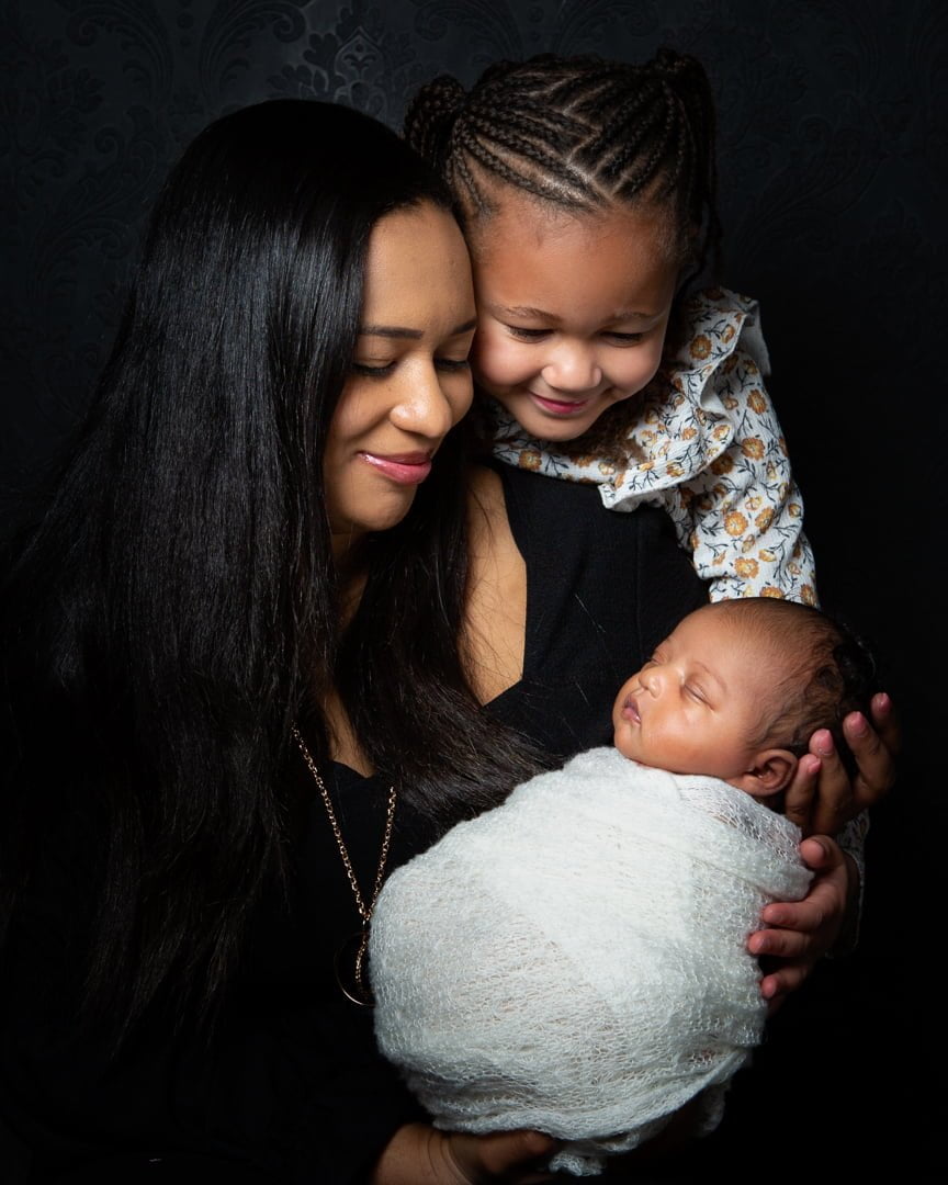 a family of a mother and her older daughter look down at a sleeping newborn wrapped in a cream swaddle