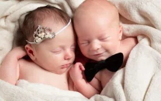 newborn twins sleep together, the girl wearing a tiny tiara and the boy with a tiny bow tie