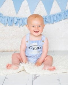 A smiling boy in a blue romper sitting in front of a wall backdrop