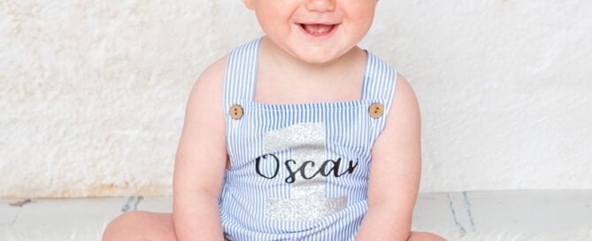 A smiling boy in a blue romper sitting in front of a wall backdrop