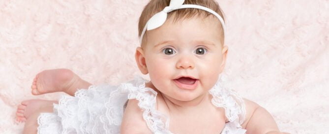 a little girl poses in a white pettiromper on her tummy on a pink blanket
