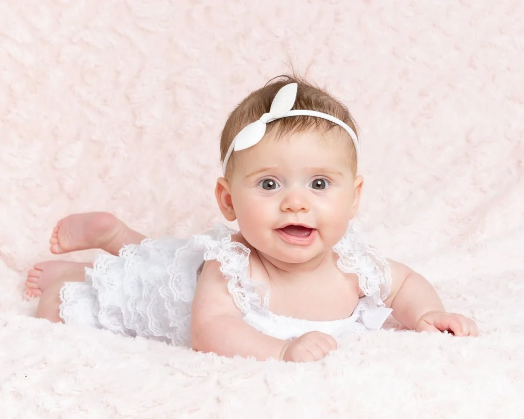 a little girl poses in a white pettiromper on her tummy on a pink blanket