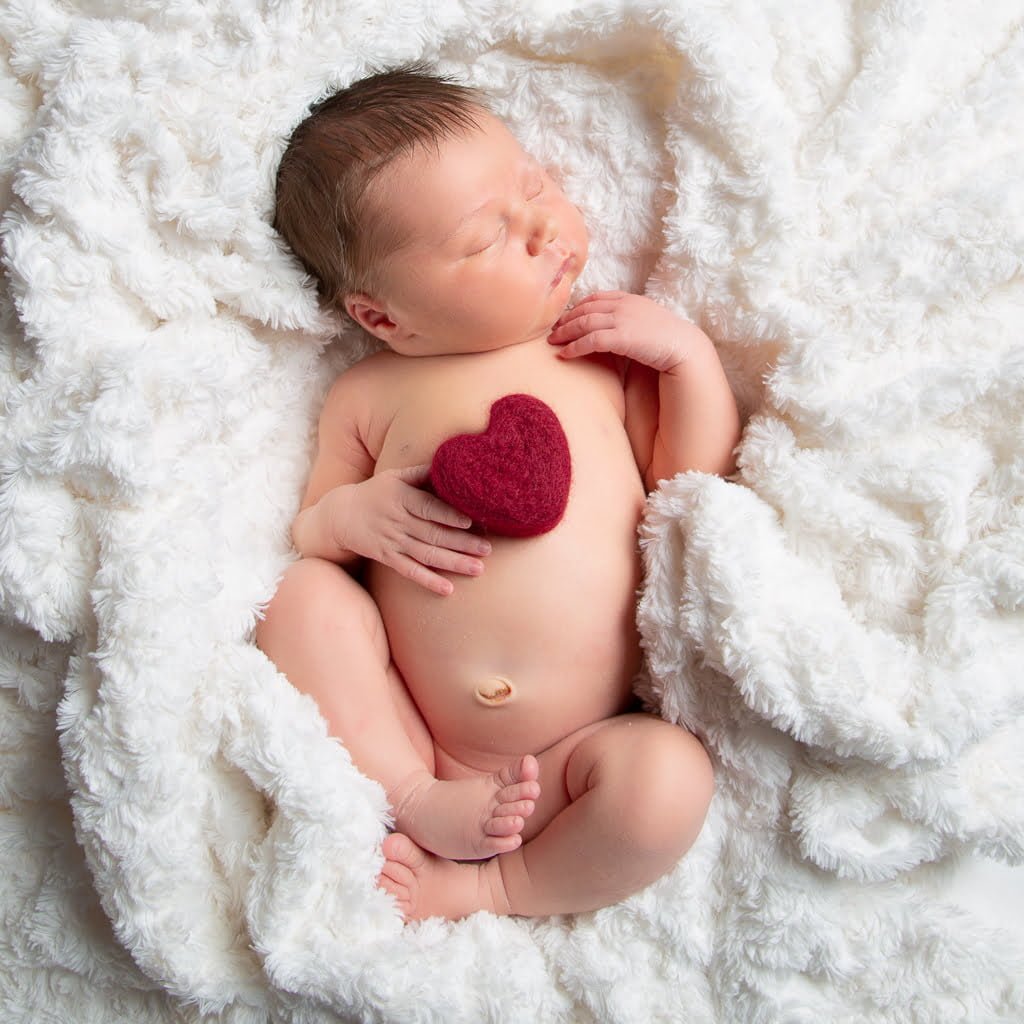 a newborn sleeping on their back on a white blankets holding a felt heart on their chest