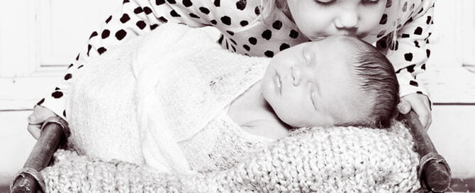 a blacka nd white image of a big sister kissing their newborn sibling