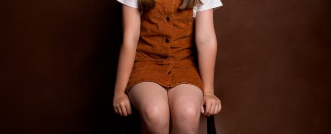 a young girl poses against a brown background wearing a dungaree dress at a fine art child portrait photo shoot