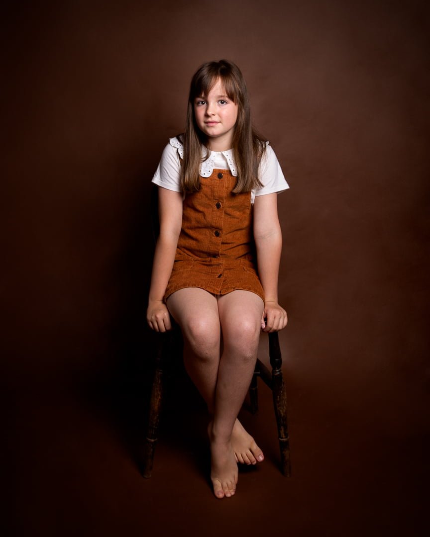 a young girl poses against a brown background wearing a dungaree dress at a fine art child portrait photo shoot