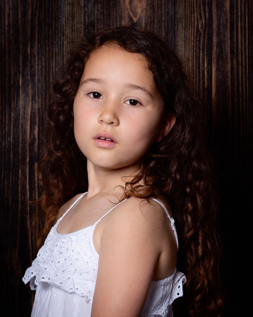 a girl poses in a white dress in front of a dark wood backdrop