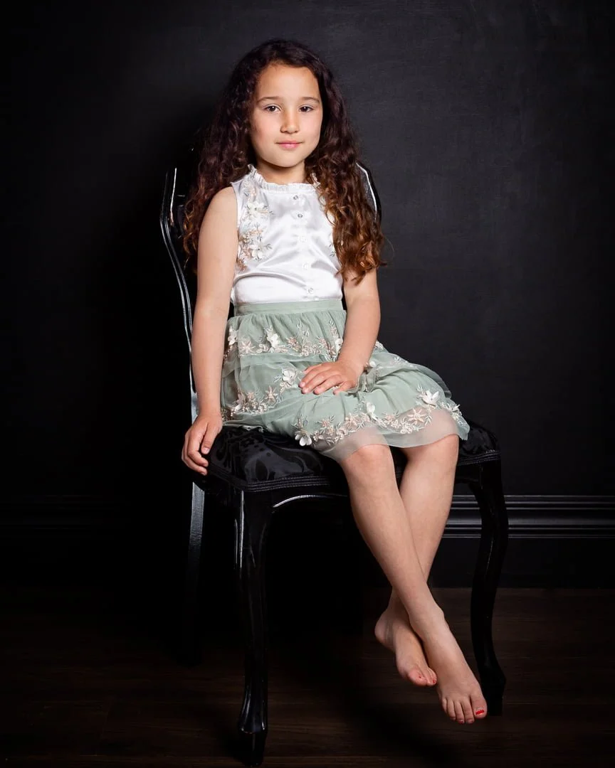 A girl sits on a black chair against a black background at a professional portrait shoot