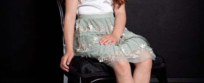 a young girl poses on a french styled chair wearing a green and white dress against a black background.