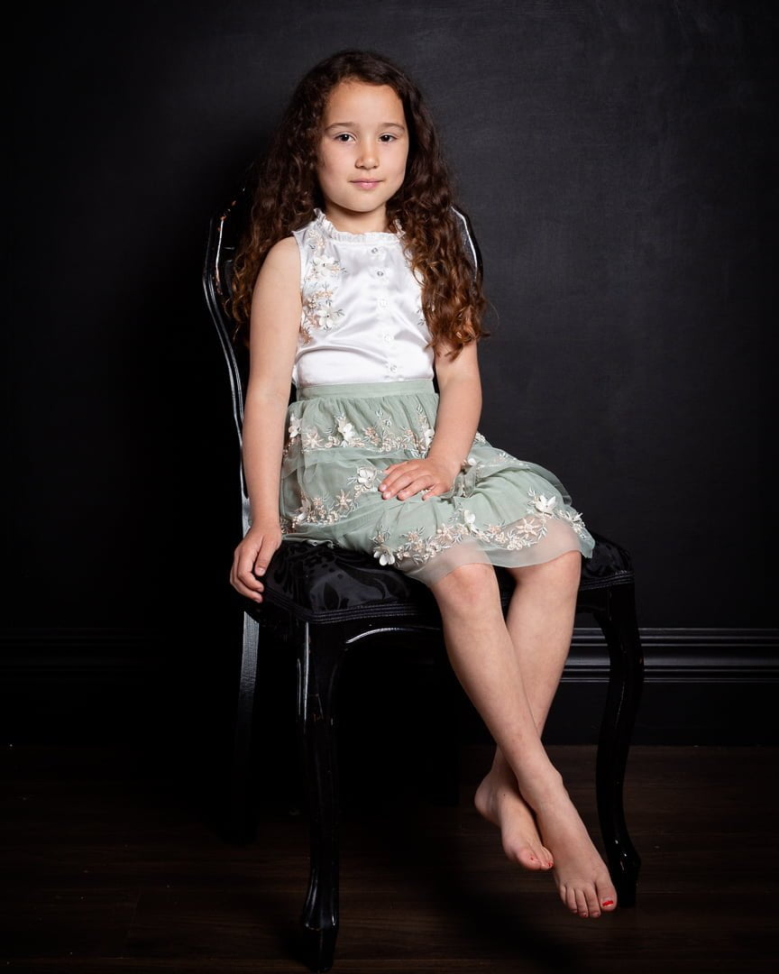 a young girl poses on a french styled chair wearing a green and white dress against a black background.