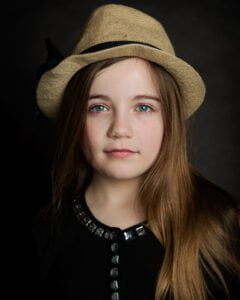 A girl poses in a hat in a classic portrait style against a dark backdrop