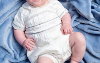 a baby in a vintage outfit looks up at ht ecamera from a blue background