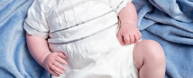 a baby in a vintage outfit looks up at ht ecamera from a blue background