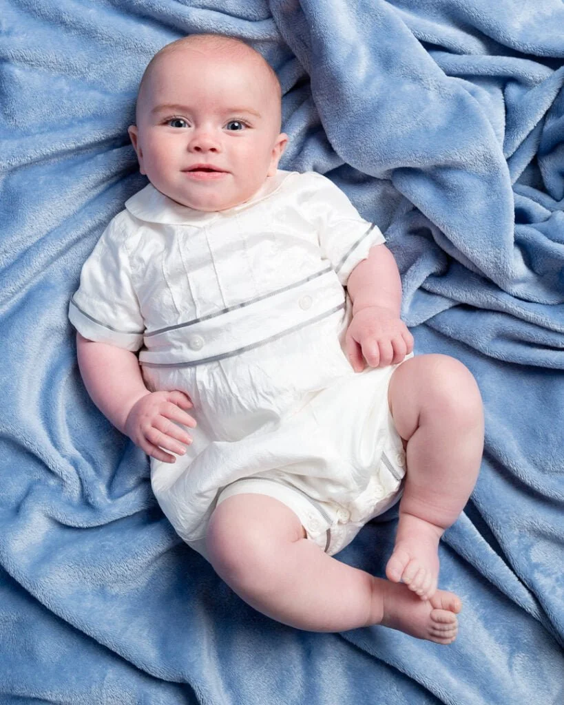 a baby in a vintage outfit looks up at ht ecamera from a blue background