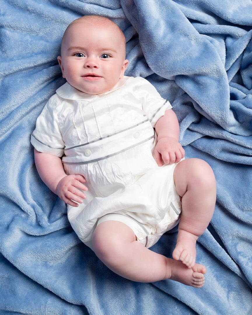 a baby in a vintage outfit looks up at ht ecamera from a blue background