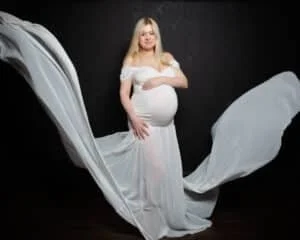 A pregnant woman in a white dress, it is tight fitting around her bump and the skirt f it is flying in the air around her.