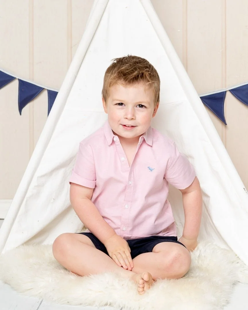 a little boy in a pink shirt poses in a miniature teepee