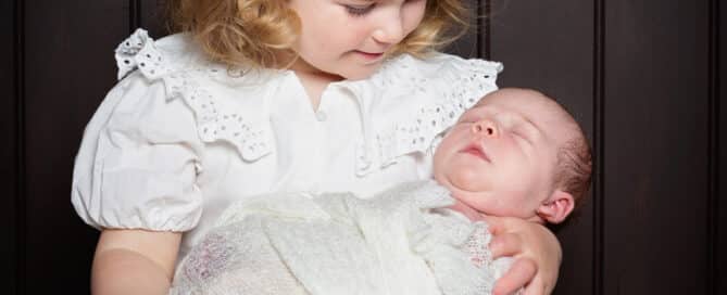 a little girl in a white dress holds her newborn sibling