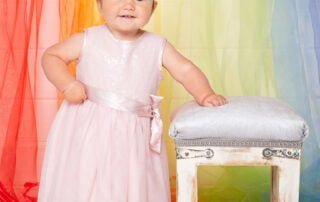a baby in a pink dress stands in front of a rainbow curtain