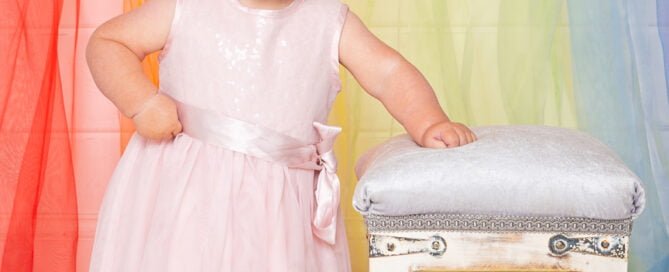 a baby in a pink dress stands in front of a rainbow curtain