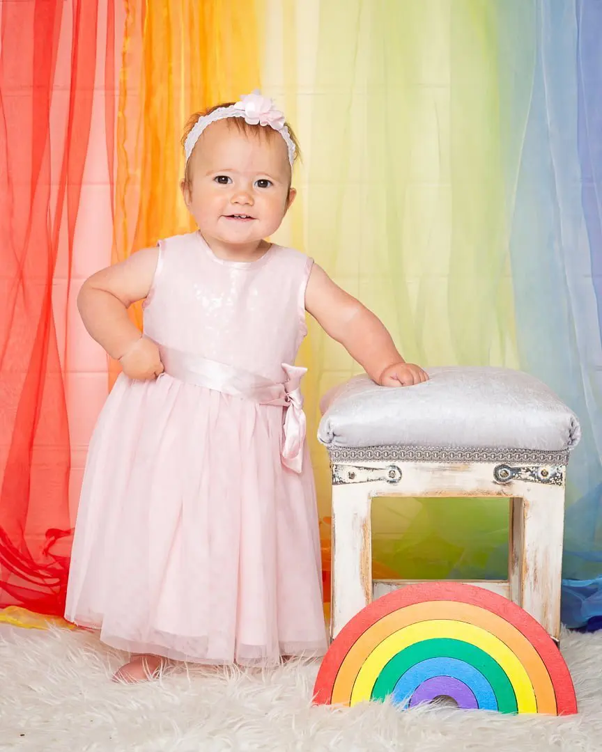 a baby in a pink dress stands in front of a rainbow curtain