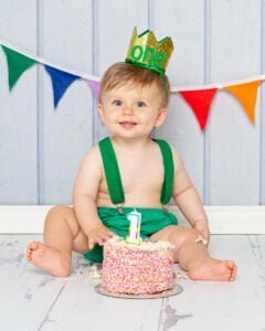  A little boy is dressed in a green and gold outfit and is smiling as he sits behind his cake at a first birthday photo shoot