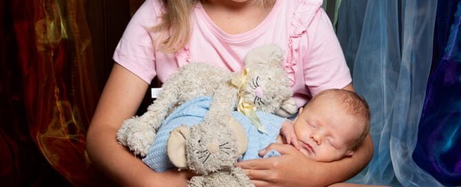 a big sister holds her baby brother against a rainbow curtain