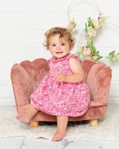  A toddler in a pink dress sits on a pink chair against a white background at a professional family photo shoot