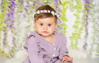 a little girl in a purple frilly outfit poses in front of a flower wall