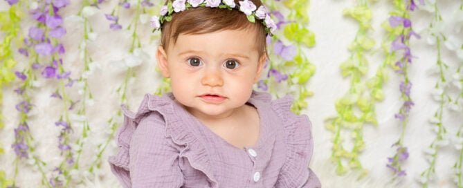a little girl in a purple frilly outfit poses in front of a flower wall