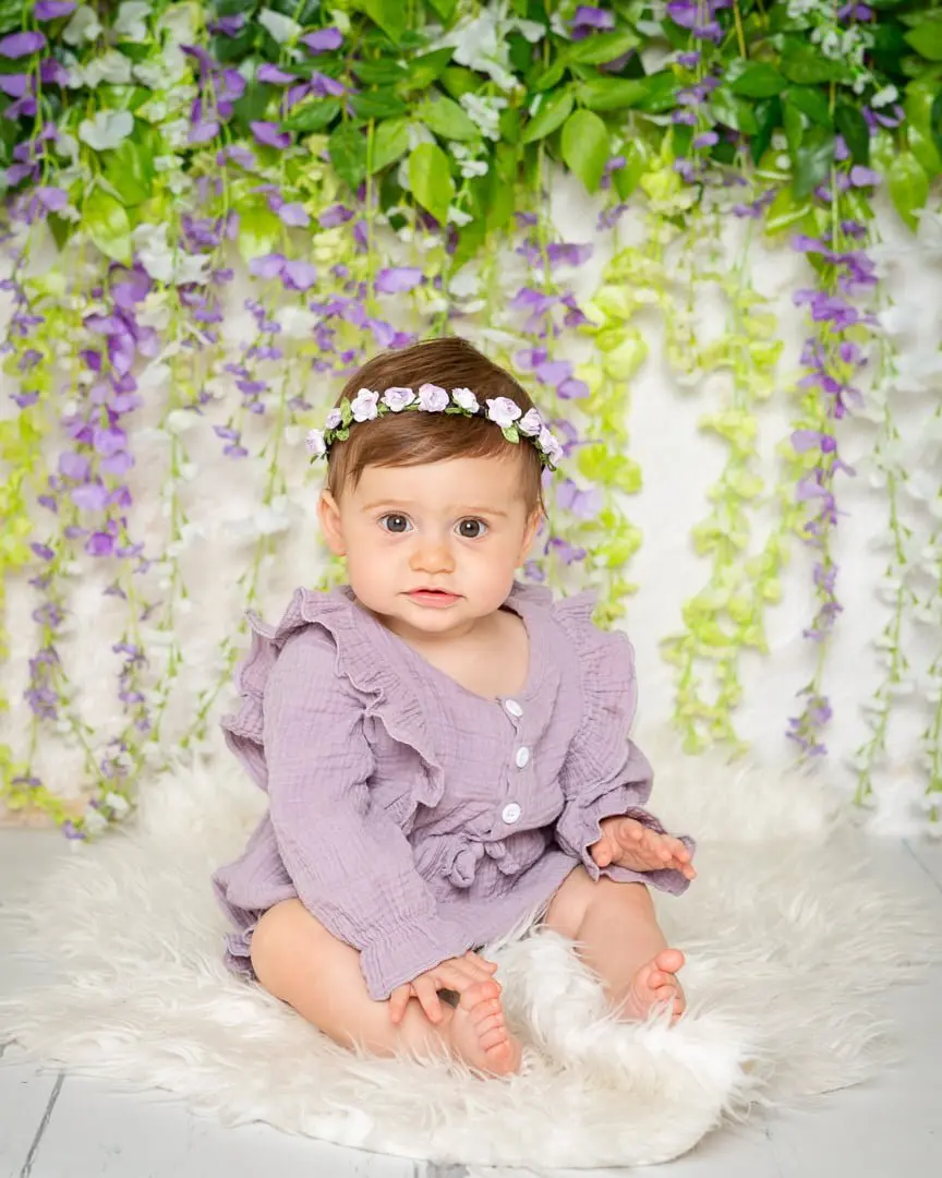 a little girl in a purple frilly outfit poses in front of a flower wall