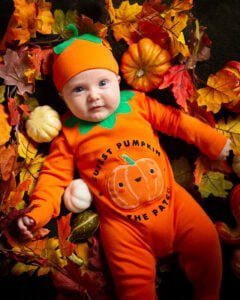  a little boy wearing a pumpkin outfit and hat looks up at the camera from a bed of mini pumpkins and leaves