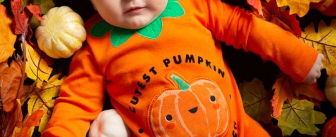 a little boy wearing a pumpkin outfit and hat looks up at the camera from a bed of mini pumpkins and leaves