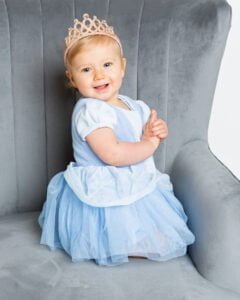  A little girl in a blue princess dress and crown sits on a grey chair and smiles for the camera at a professional baby photo shoot