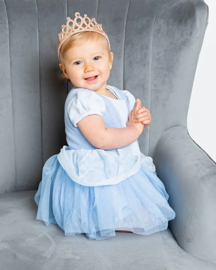 Baby Photography in East Grinstead West Sussex alittle girl in a princess outfit smiles on a grey chair