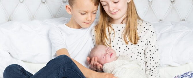 two older siblings hold their newborn sibling on a bed