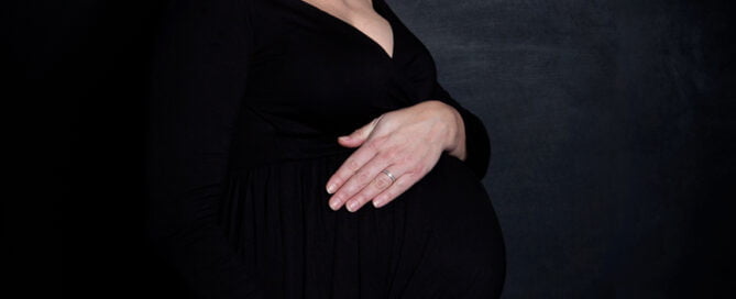 A pregnant woman in a black dress is looking down towards her bump against a black background at a professional maternity photo shoot