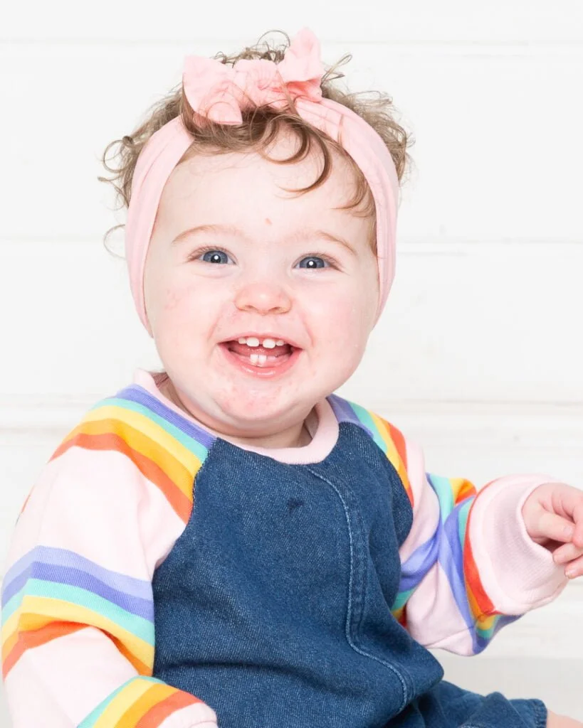 A baby in a denim and rainbow outfit with spots on their face edited out