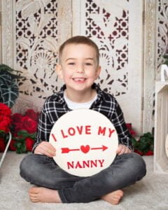 a little boy sits in front of a screen holding a sign saying I love my Nanny