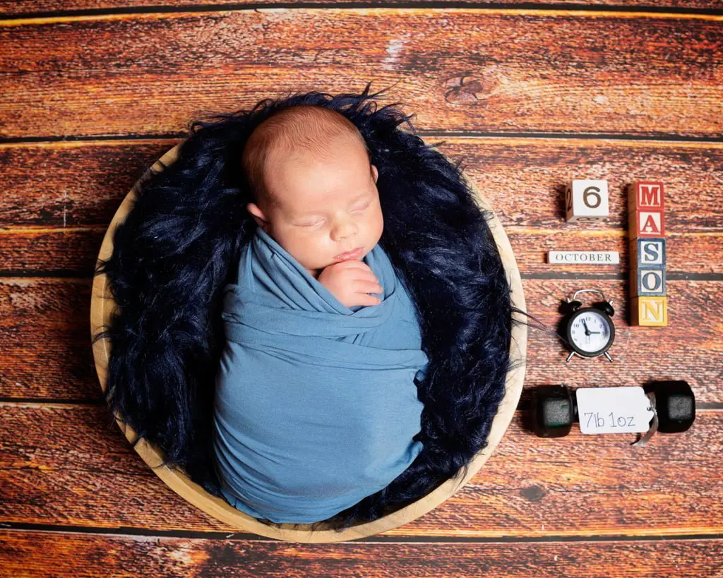 a newborn with a birth announcement prop set in a bowl wrapped in blue
