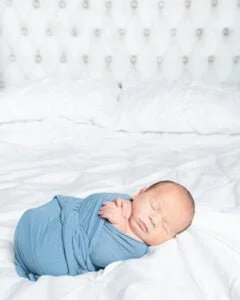 a newborn sleeping on a white bed swaddles in a blue wrap