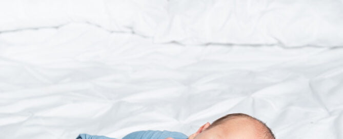 a newborn sleeping on a white bed swaddles in a blue wrap