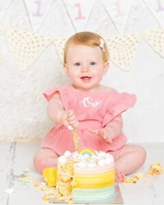 A little girl is wearing a pink dress and sitting behind a birthday cake which she is enjoying at a first birthday professional photo shoot