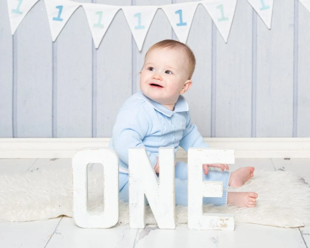 a little boy is dressed in a blue romper and sits smiling behind some wooden letters that spell out ONE